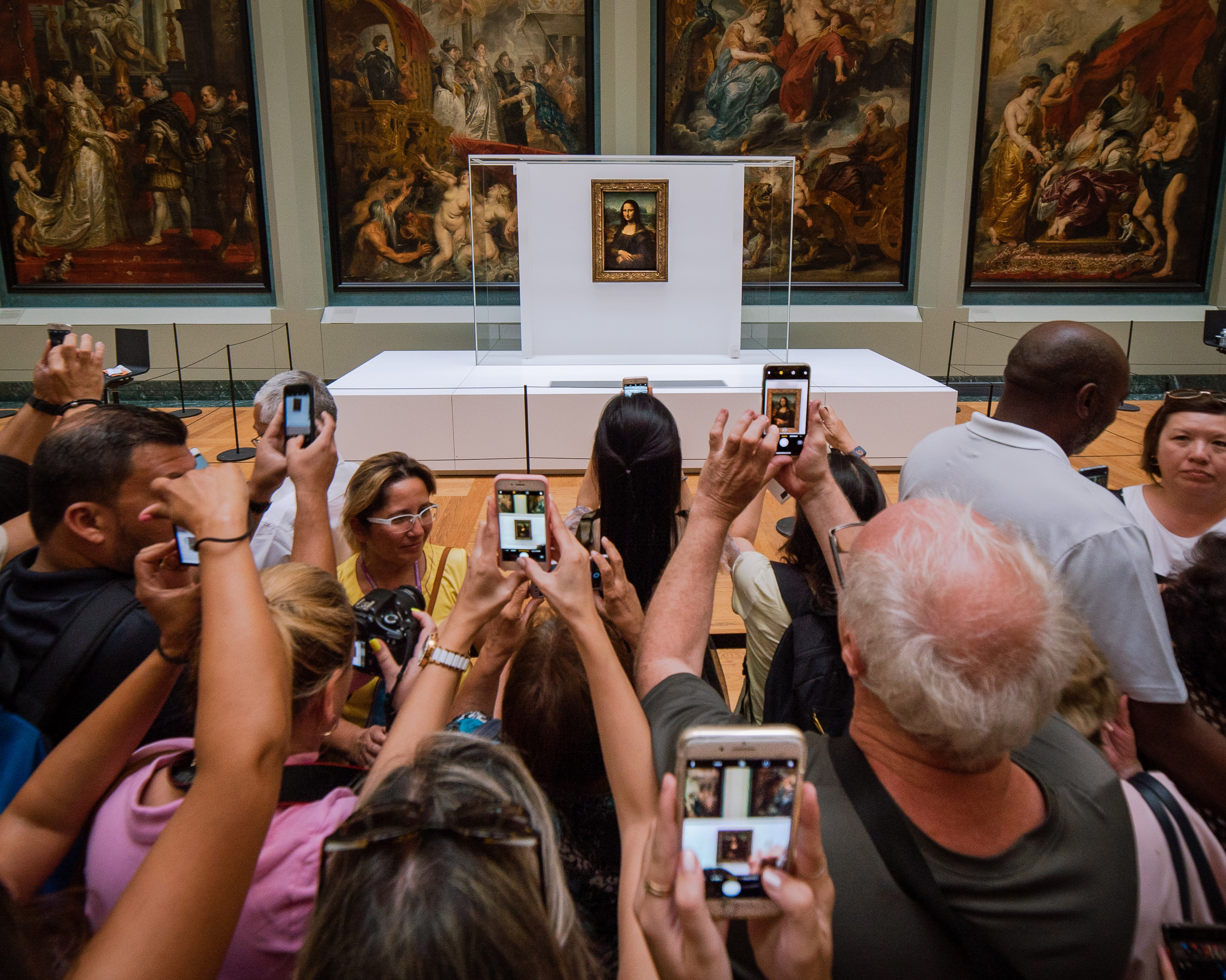 A large crowd is gathered around the Mona Lisa painting holding up phone cameras taking pictures.