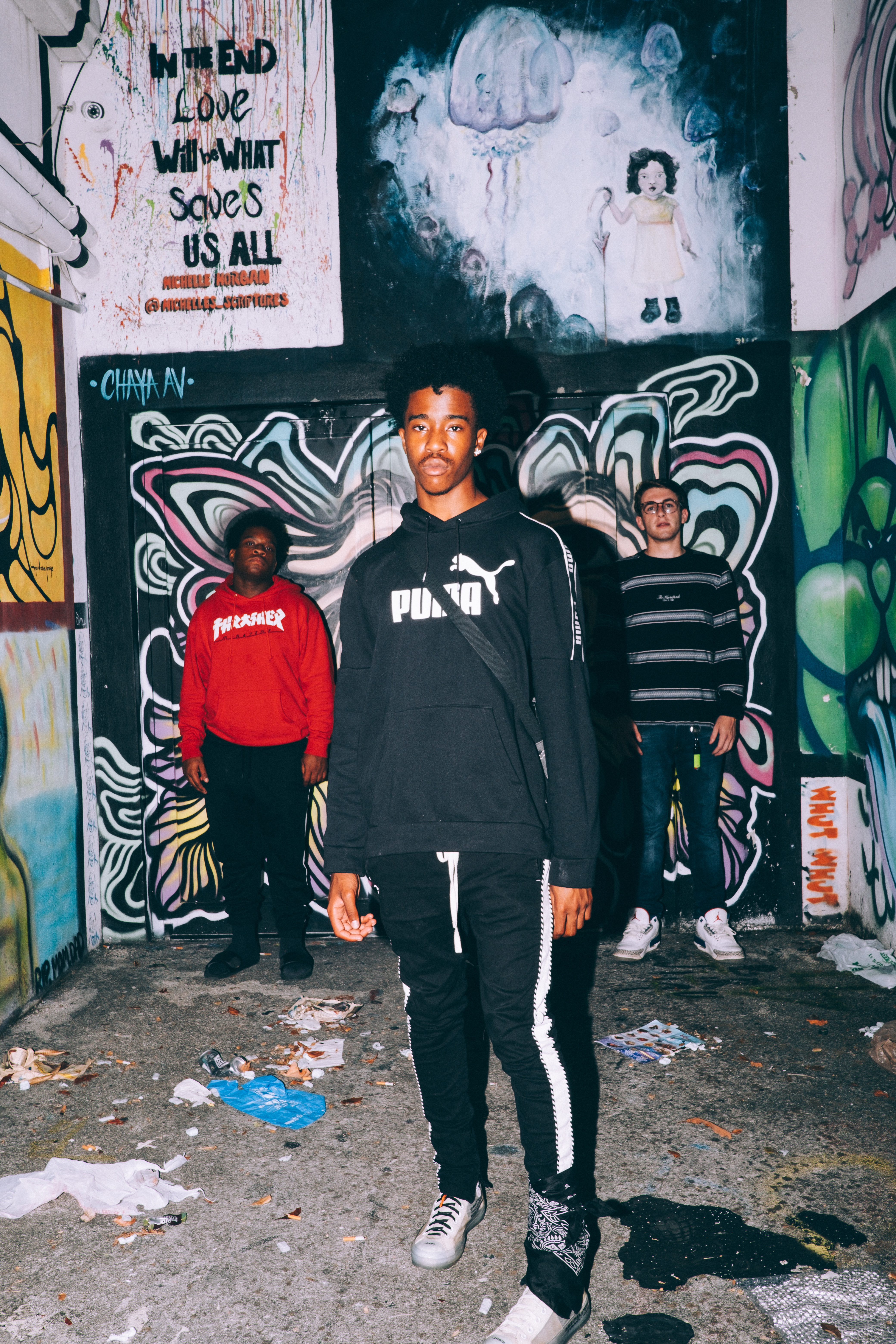 Three men staggered in a photograph. One man in all black is closer to the camera, with the two on each side of his standing farther back. The are surrounded by concrete walls with graffiti and there is trash on the ground.