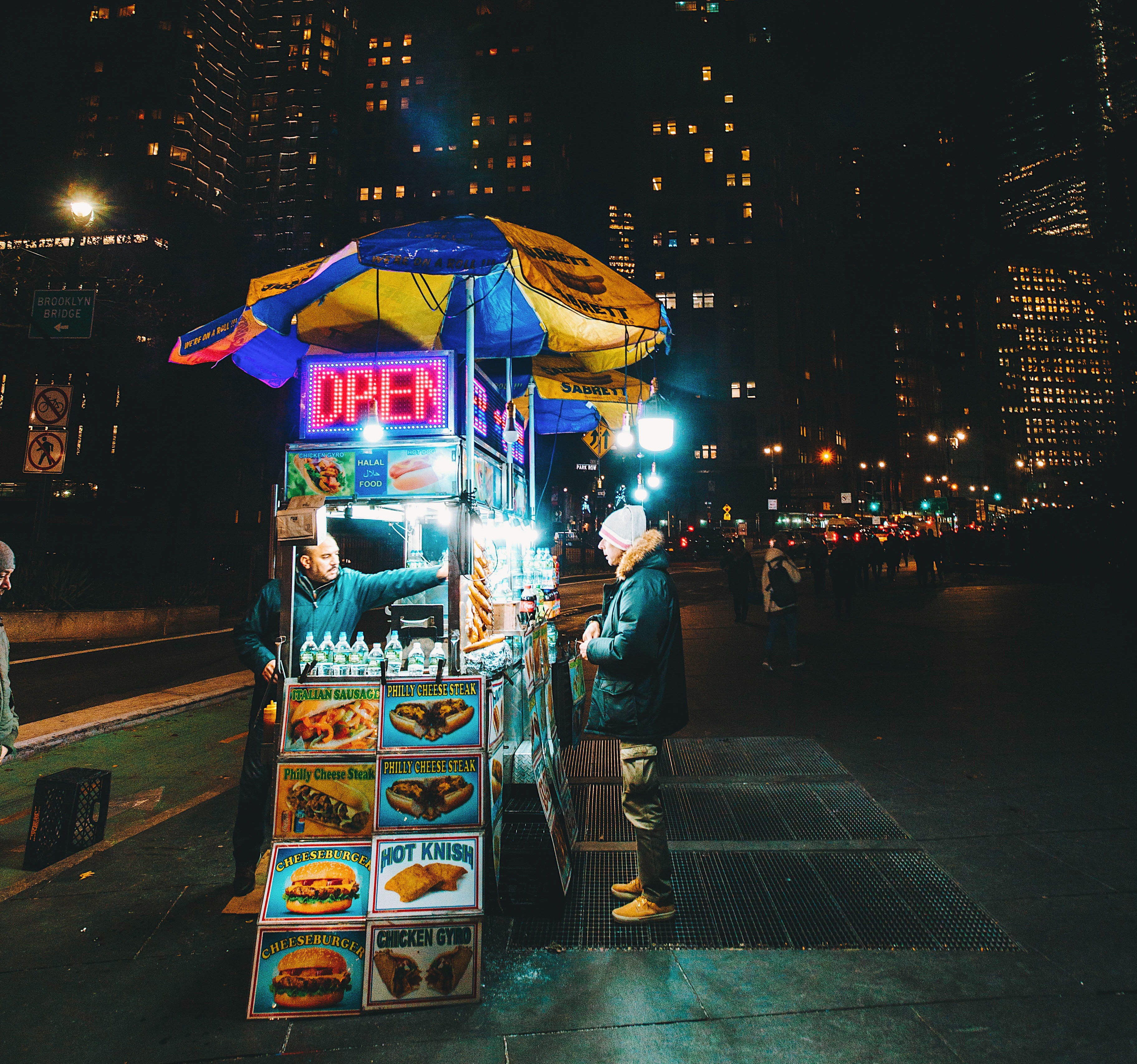Food cart with an open sign where a man is buying food and New York City skyline is in the background.